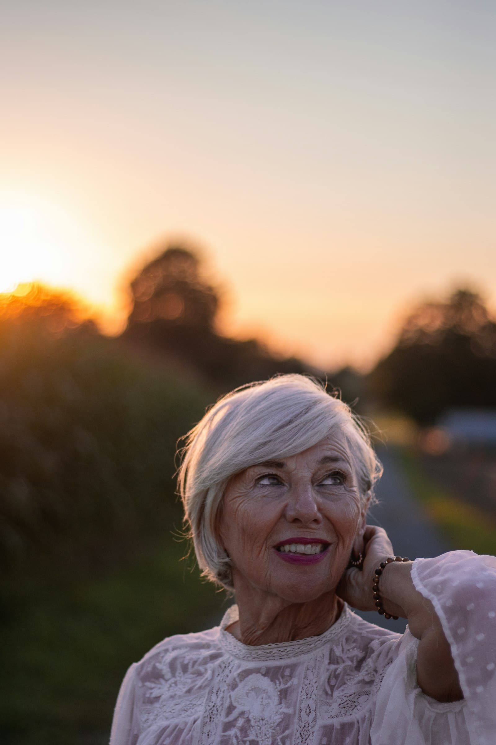 A woman with short white hair smiles and looks up, hand at her ear, at sunset.