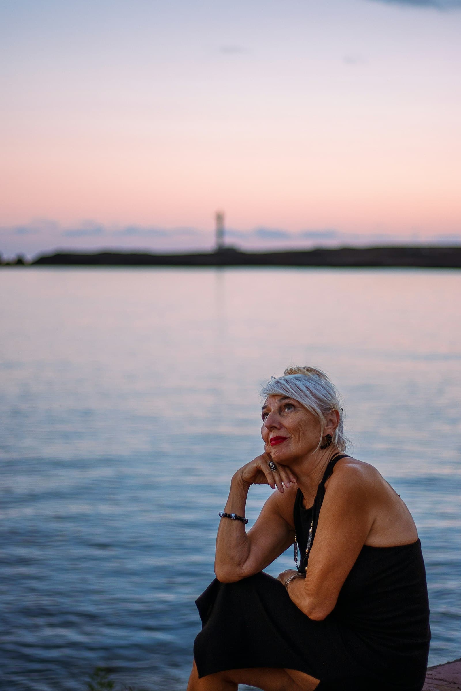 A woman with short silver hair sits by the water at dusk, looking up at a pastel sky.