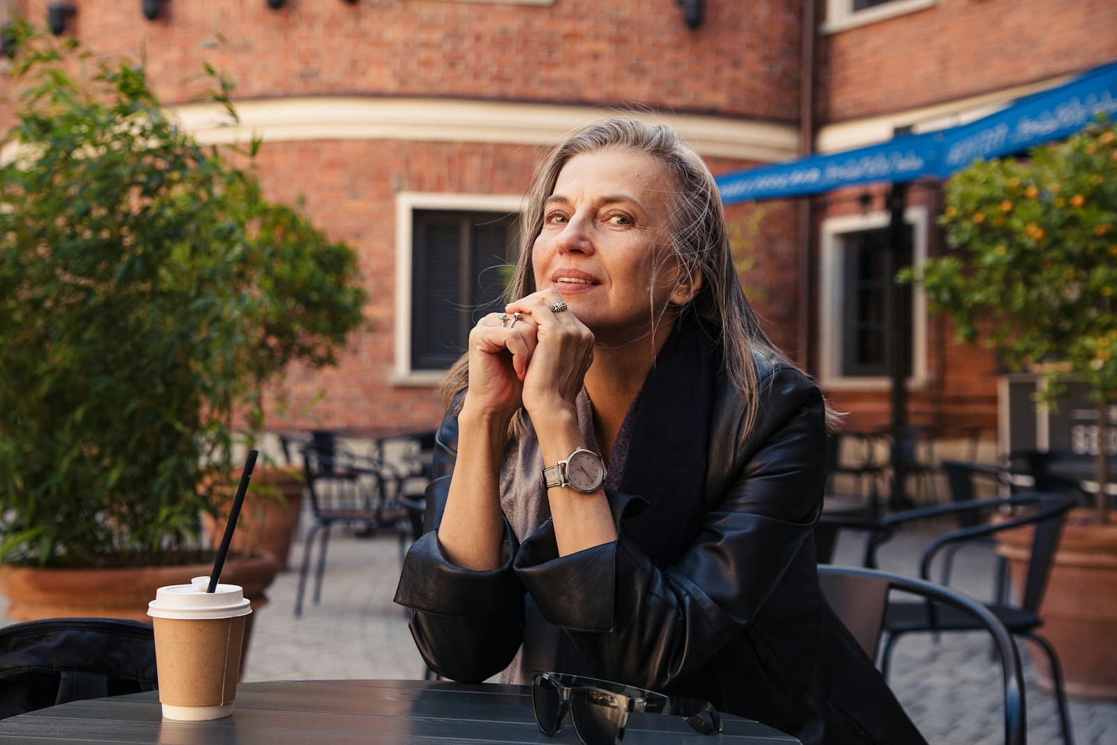 A woman with long grey hair sits at a cafe table outdoors, chin resting on her hands, smiling.